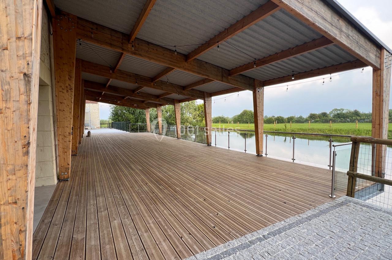 Terrasse en bois couverte avec vue sur un plan d'eau et des espaces verts, entourée de garde-corps en métal et verre.