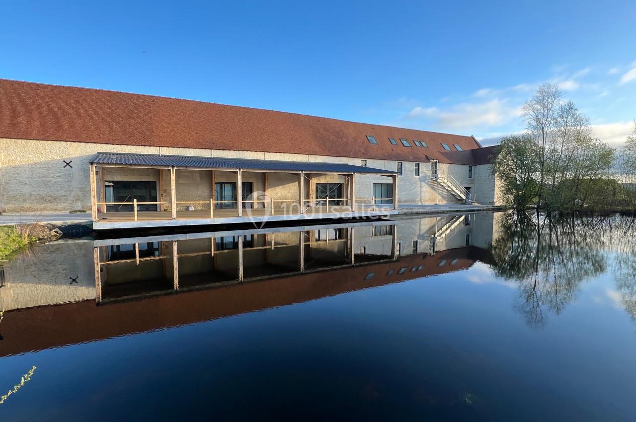 Bâtiment en pierre avec toit rouge, grandes baies vitrées et terrasse en bois, reflété dans un plan d'eau calme.