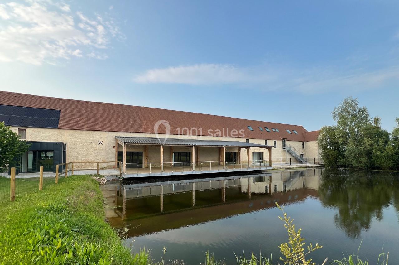 Bâtiment moderne en pierre avec toit en pente, bordé par un étang et entouré de végétation sous un ciel dégagé.