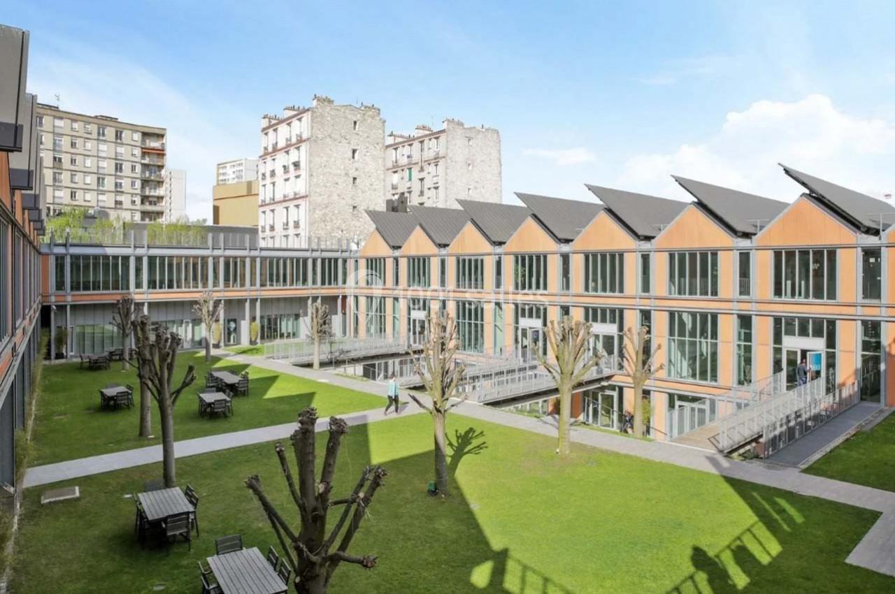 Cour intérieure avec pelouse, arbres taillés, tables, et bâtiments modernes en verre et bois sous un ciel dégagé.