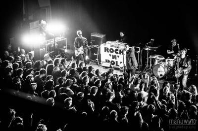 Un technicien supervise un enregistrement en studio, avec trois personnes chantant derrière une vitre insonorisée.