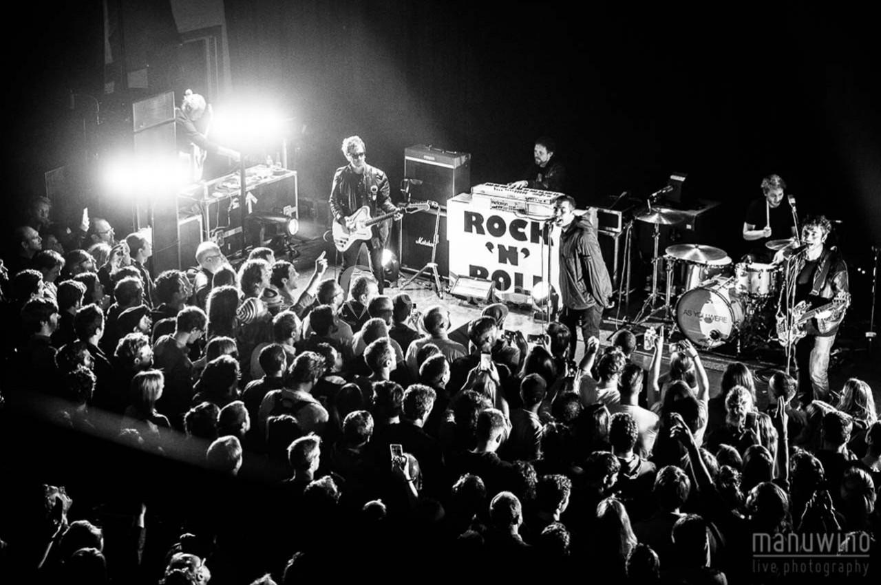 Groupe de musiciens jouant sur scène devant une foule dans une ambiance de concert en noir et blanc.