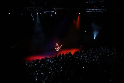 Un technicien supervise un enregistrement en studio, avec trois personnes chantant derrière une vitre insonorisée.