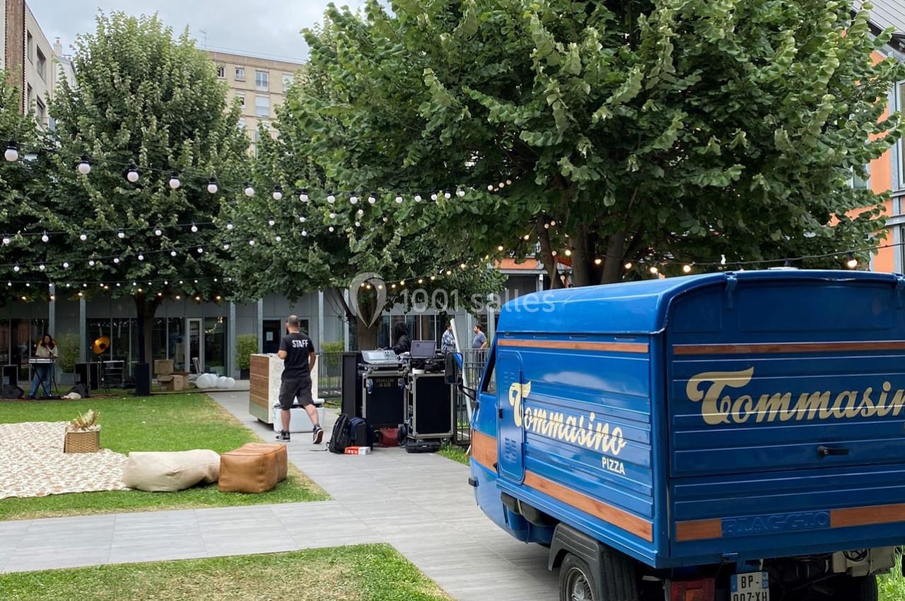 Un triporteur bleu stationné dans une cour arborée avec des équipements techniques et des guirlandes lumineuses suspendues.