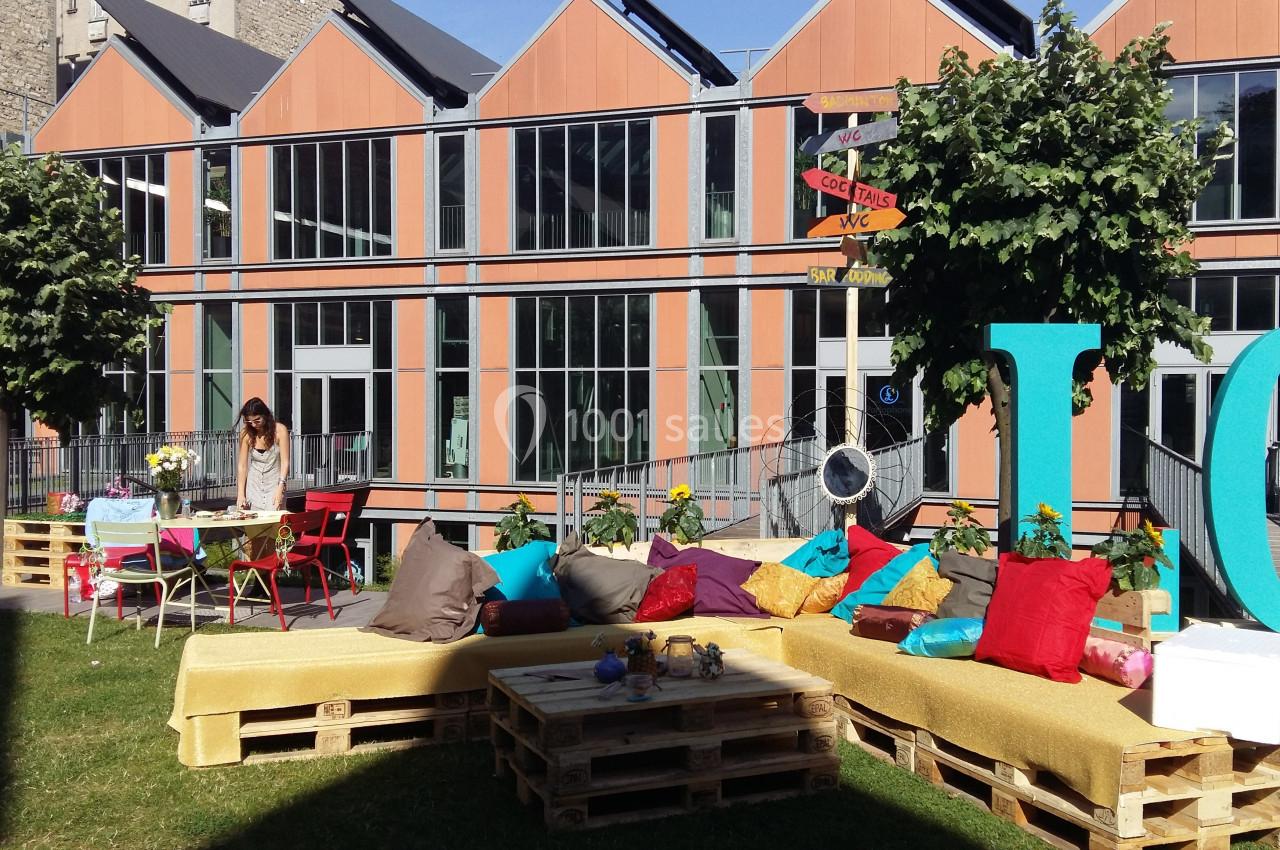 Espace extérieur aménagé avec des palettes, coussins colorés et tables, devant un bâtiment moderne avec grandes fenêtres.