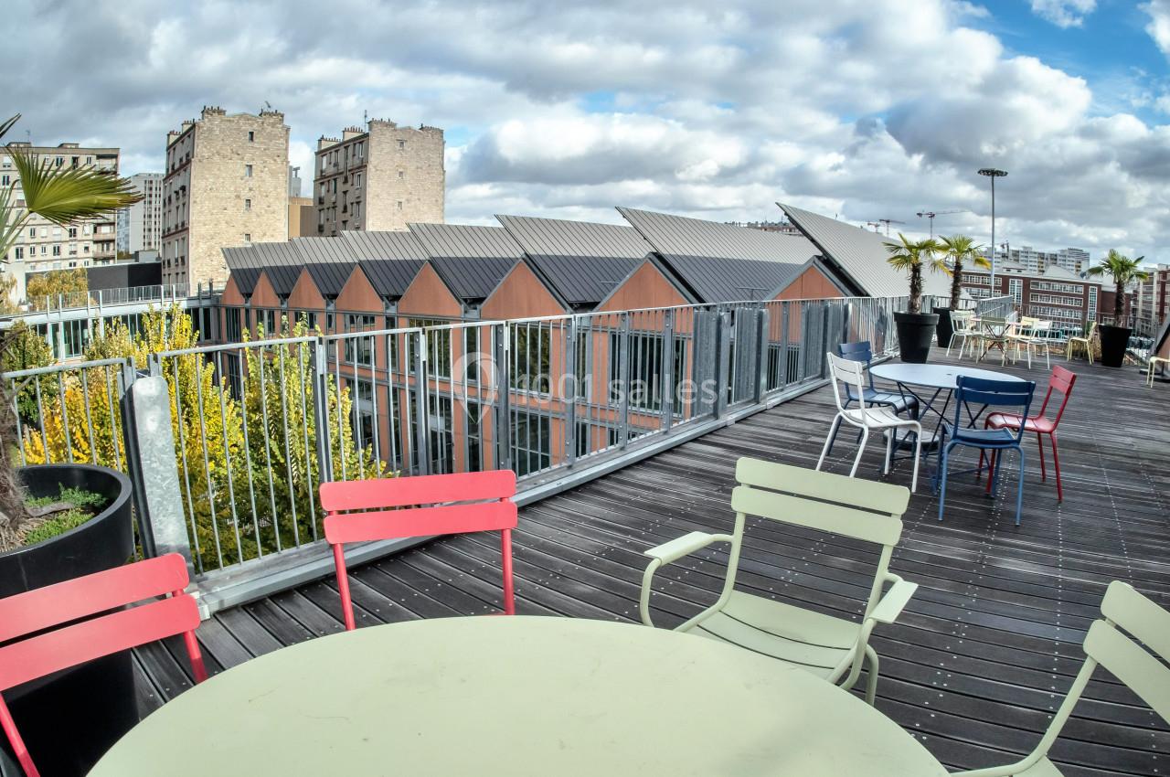 Terrasse en bois avec tables et chaises colorées, vue sur des toits en pente et des bâtiments urbains sous un ciel nuageux.