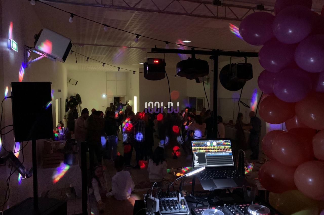 Salle de fête sombre avec des lumières colorées, une table de mixage au premier plan et des invités dansant en arrière-plan.