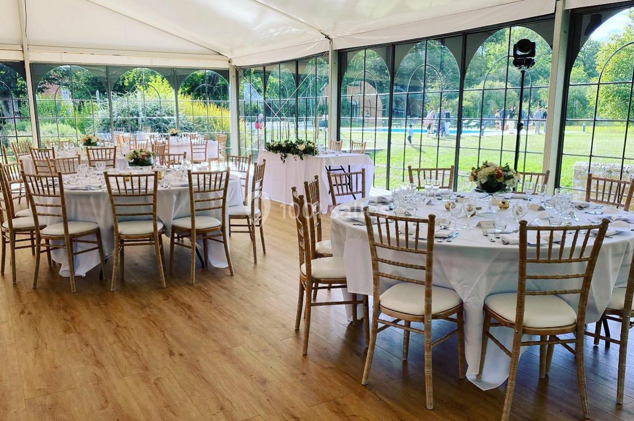 Salle de réception sous une tente avec tables rondes dressées, chaises en bois clair et vue sur un jardin verdoyant.
