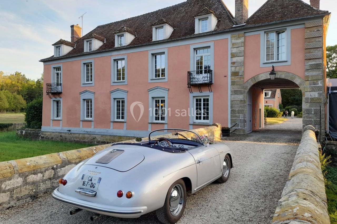 Voiture ancienne décapotable argentée stationnée devant une grande maison de style classique avec façade rose.
