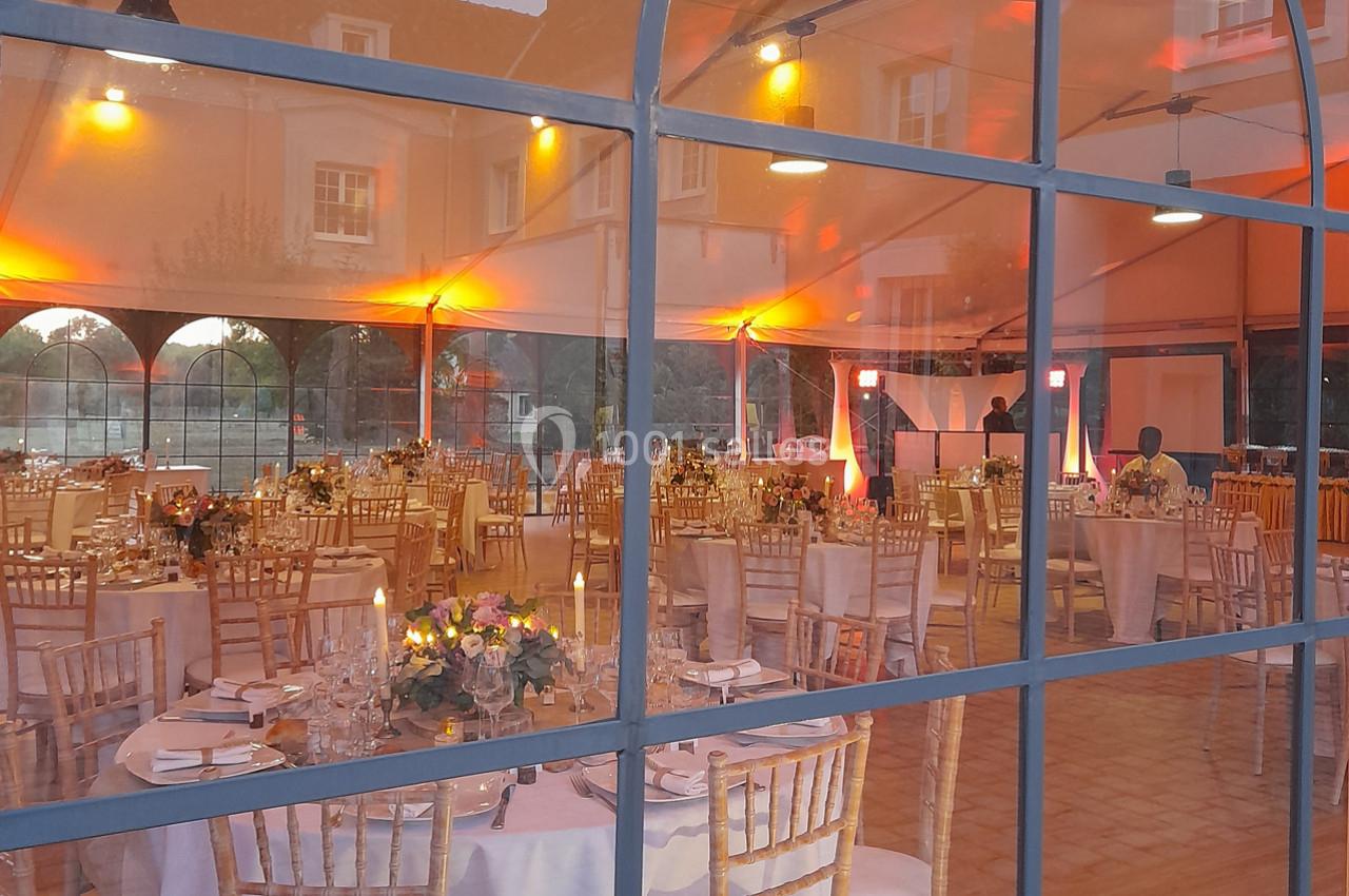 Salle de réception décorée avec des tables dressées, des chaises en bois et un éclairage chaleureux visible à travers une…