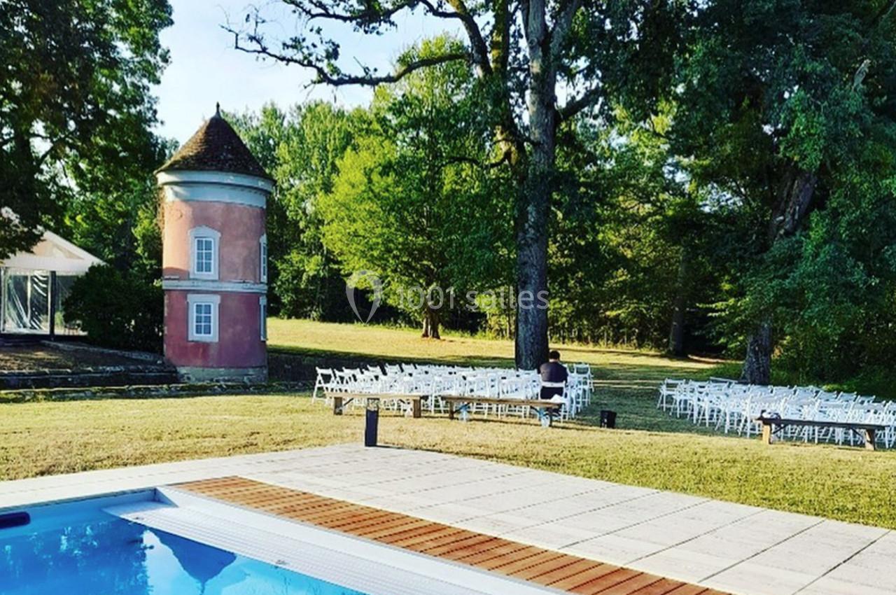 Vue d'une piscine entourée de dalles, avec un petit bâtiment rose, des arbres et des chaises blanches en arrière-plan.
