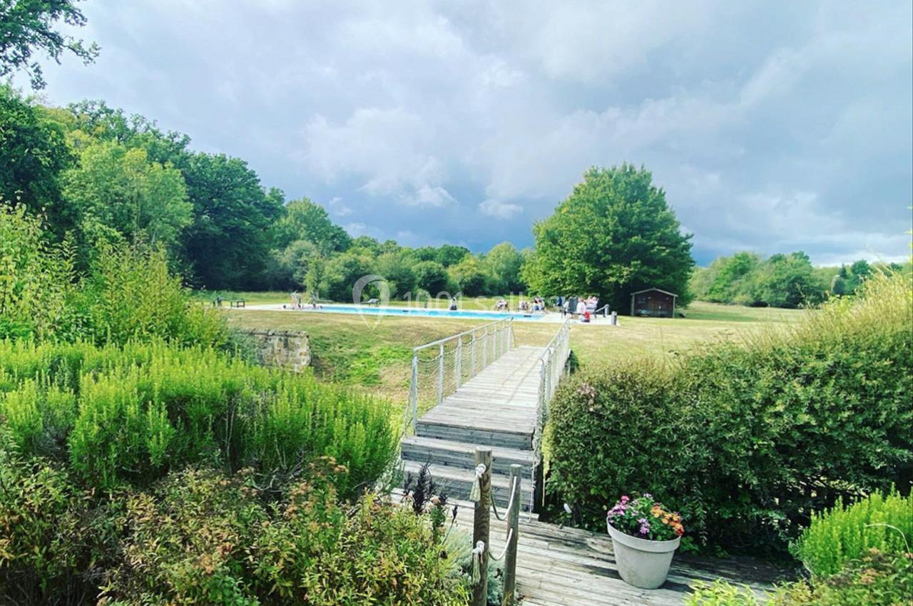 Passerelle en bois traversant un jardin verdoyant, menant à une prairie avec une piscine et des arbres en arrière-plan.