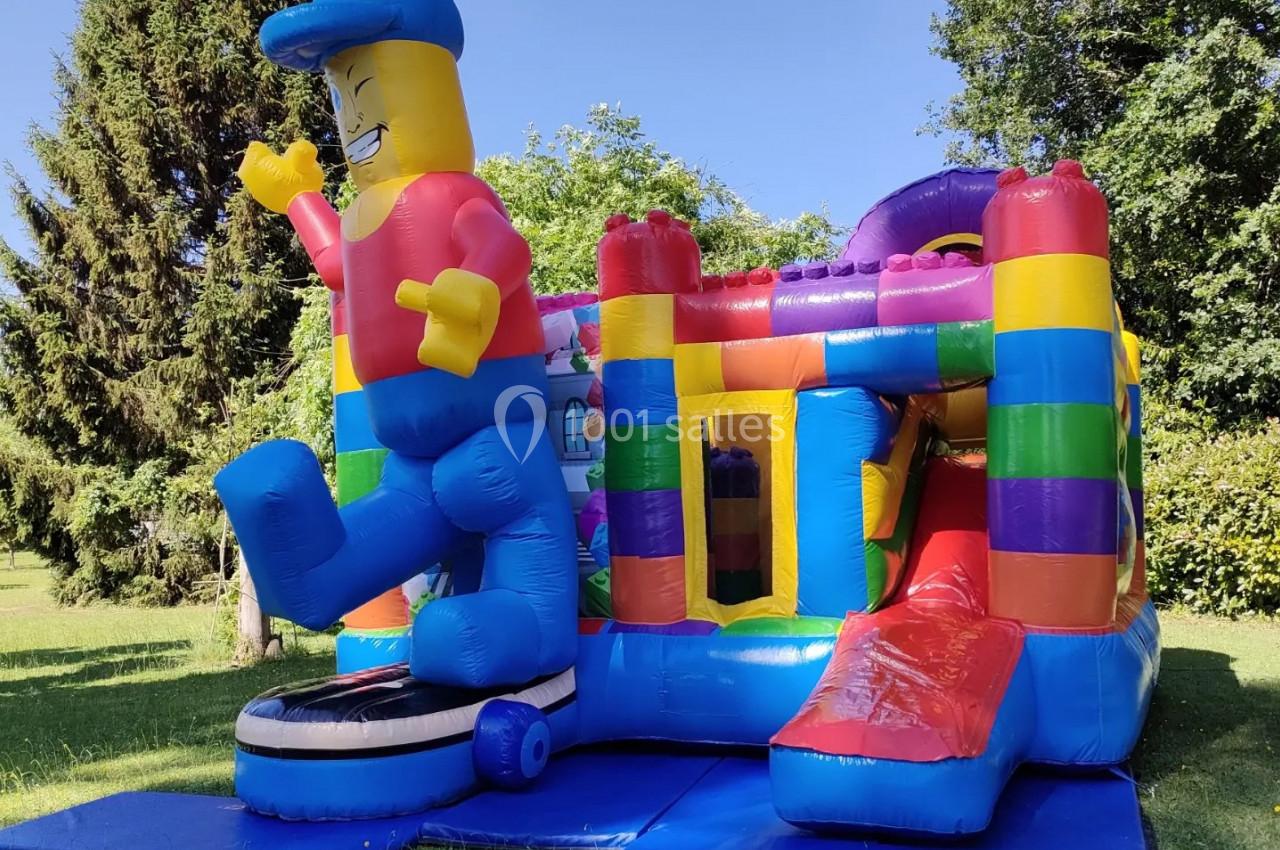 Structure gonflable colorée en plein air avec toboggan et personnage souriant, entourée de verdure.