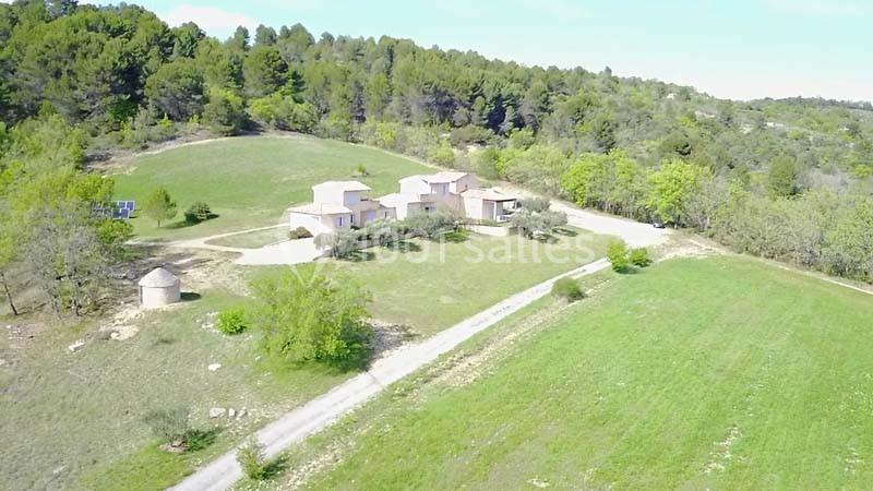 Vue aérienne d'une maison entourée de champs verdoyants et de collines boisées sous un ciel dégagé.