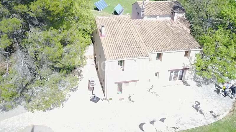 Vue aérienne d'une maison en pierre avec toit en tuiles, entourée d'arbres et de panneaux solaires au sol.