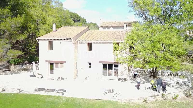 Vue aérienne d'une maison en pierre avec terrasse, entourée d'arbres et d'un jardin spacieux par temps ensoleillé.