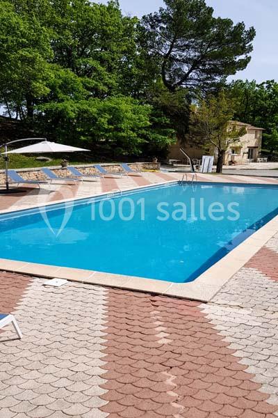 Piscine extérieure entourée de chaises longues, parasols et arbres, avec une maison en arrière-plan.