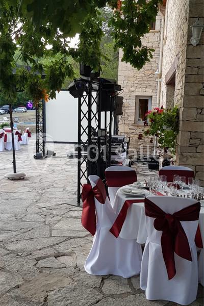 Tables de réception décorées de nappes blanches et nœuds rouges, installées en extérieur près d'un bâtiment en pierre.