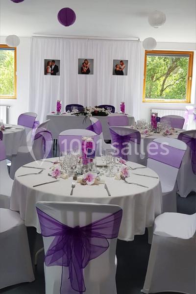 Salle décorée pour un événement avec nappes blanches, chaises ornées de nœuds violets et tables dressées.