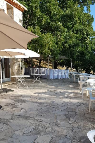 Terrasse en pierre avec tables et chaises blanches, parasols beiges et arbres en arrière-plan.