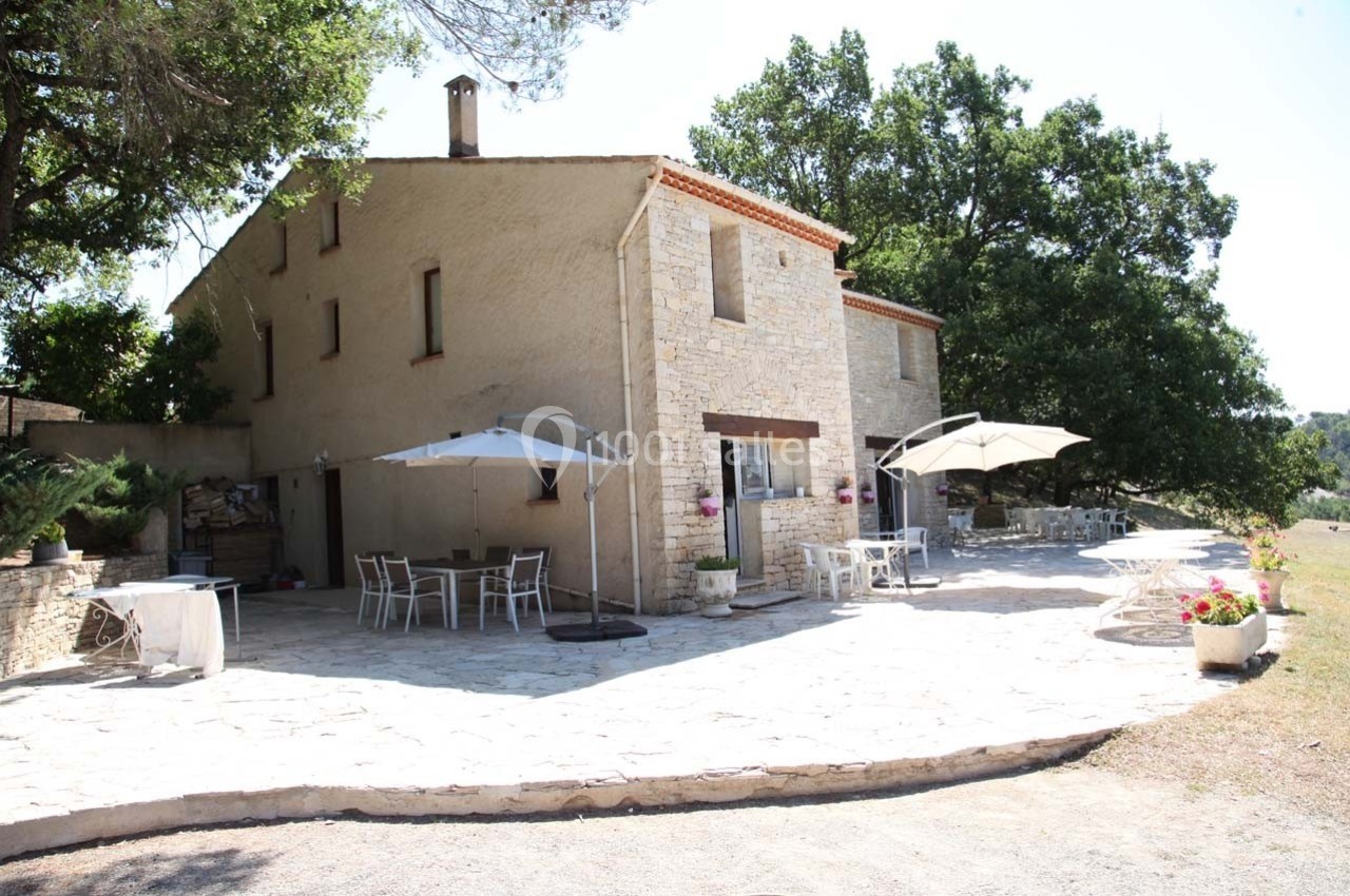 Maison en pierre avec terrasse aménagée, tables, chaises et parasols, entourée d'arbres et d'un espace naturel.