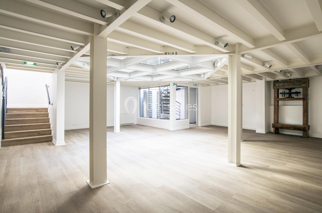 Salle lumineuse avec parquet clair, poutres apparentes blanches, escalier à gauche et grande verrière au plafond.