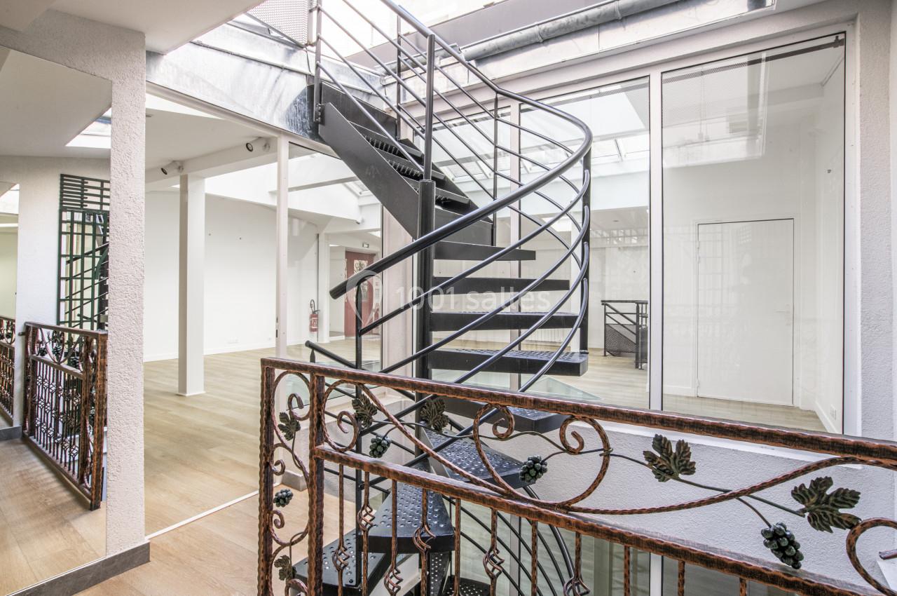 Escalier en colimaçon noir dans un espace lumineux avec parquet, verrières et garde-corps décoratif en métal.