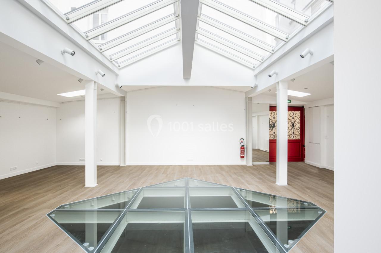 Espace intérieur lumineux avec verrière, parquet clair et une structure en verre au sol, porte rouge en arrière-plan.