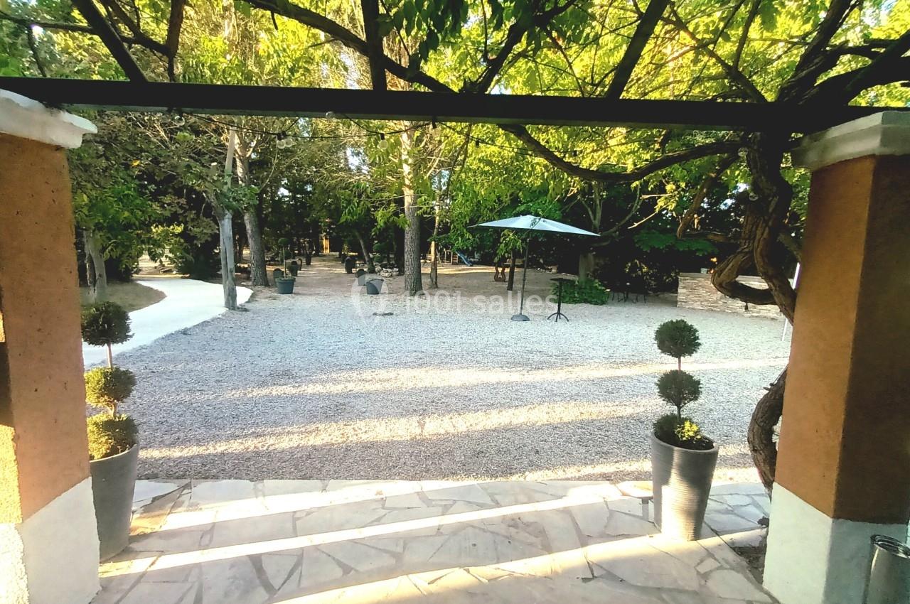 Vue d'une cour extérieure avec sol en gravier, arbres ombrageants et parasol, encadrée par une arche en pierre.