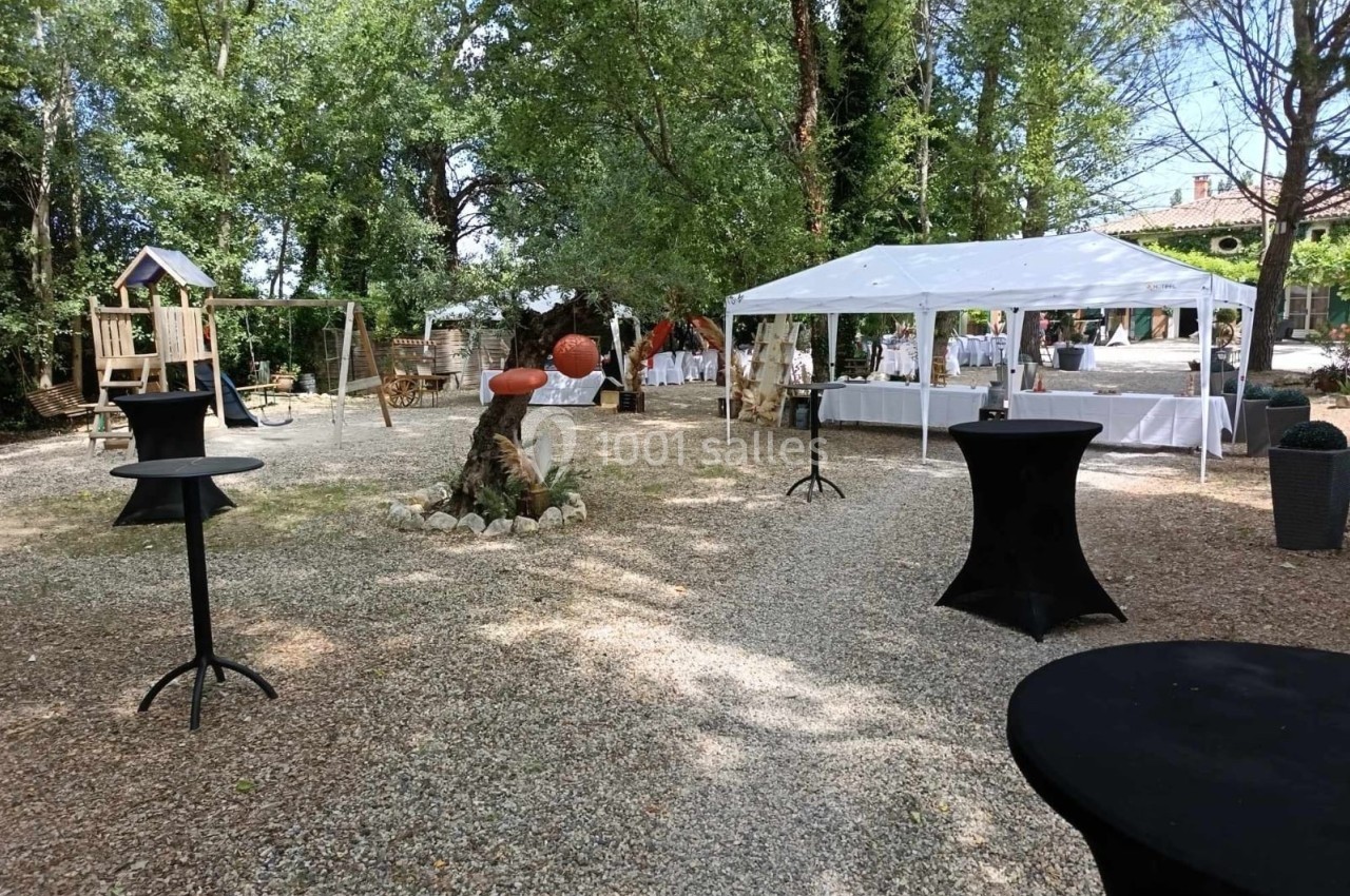 Espace extérieur avec tables sous un chapiteau blanc, aire de jeux en bois et décoration naturelle sur sol gravillonné.