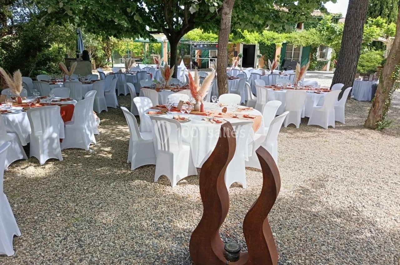 Tables décorées pour un événement en plein air, disposées sous des arbres, avec nappes blanches et ornements naturels.