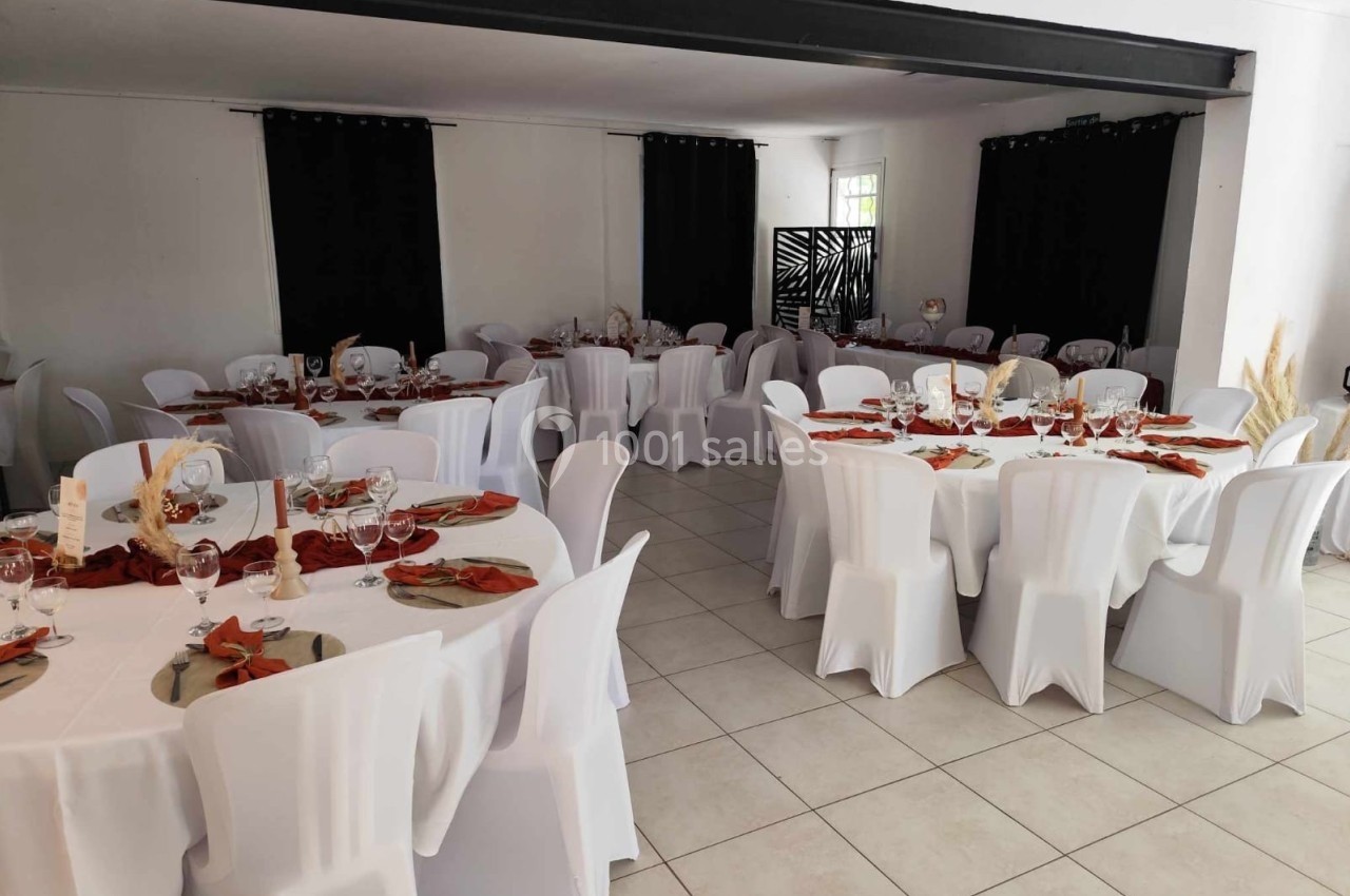 Salle de réception décorée avec des tables rondes, nappes blanches, serviettes rouges et chaises recouvertes.