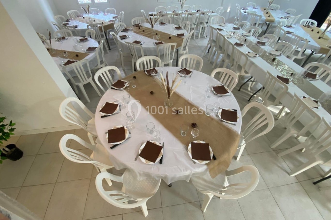 Salle décorée pour un événement, avec des tables rondes dressées, nappes blanches et chemins de table en toile de jute.