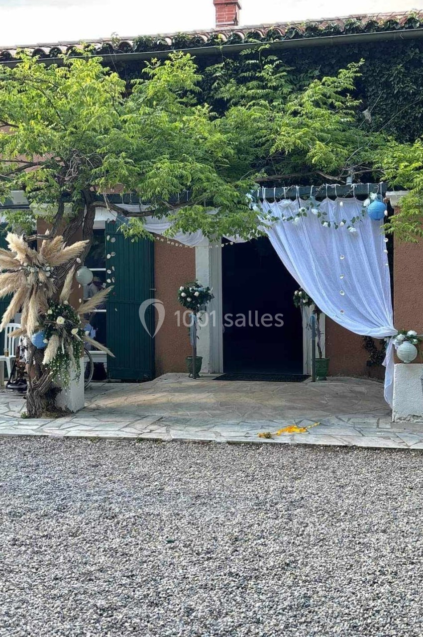 Entrée décorée avec rideaux blancs, guirlandes lumineuses, plantes et ornements floraux devant un bâtiment rustique.