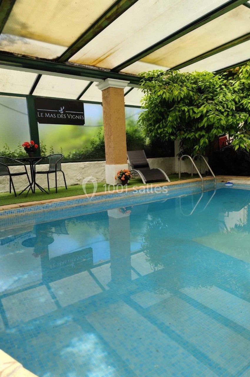 Piscine intérieure avec chaises longues, table et plantes, sous une verrière partiellement ombragée.