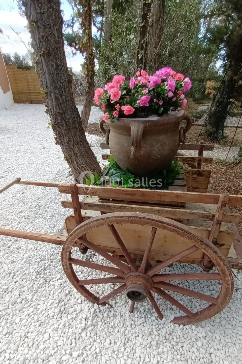 Charrette en bois ancien avec une grande jarre en terre cuite remplie de fleurs roses, posée sur un sol de gravier.