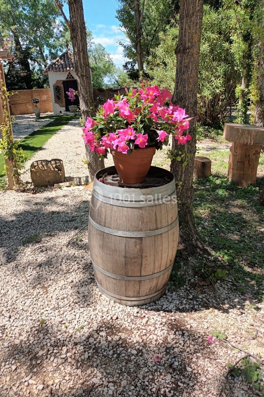 Pot de fleurs roses posé sur un tonneau en bois, dans un jardin ensoleillé entouré d'arbres et de mobilier rustique.
