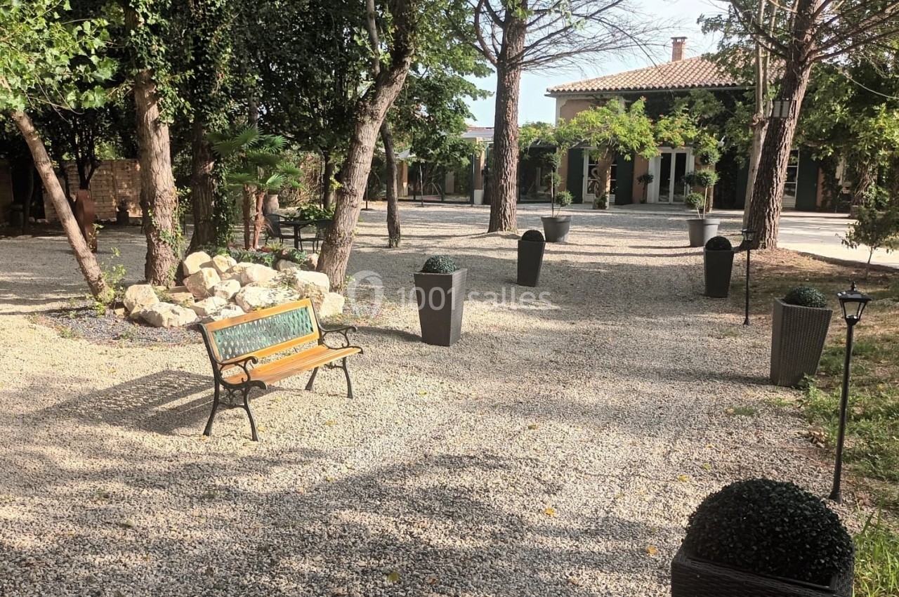 Allée gravillonnée bordée d'arbres, avec banc en bois, pots décoratifs et maison en arrière-plan.