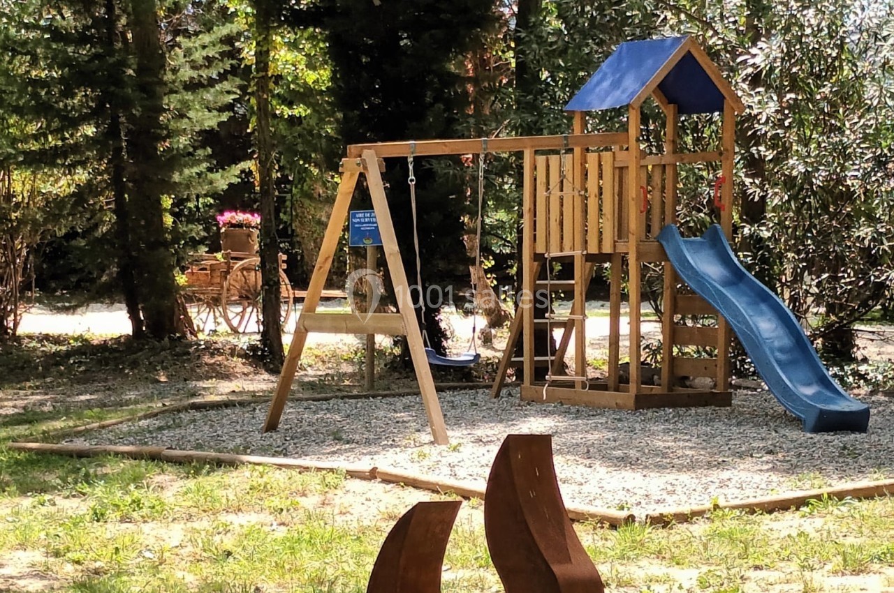 Aire de jeux en bois avec toboggan bleu, balançoire et échelle, entourée d'arbres et de gravier.