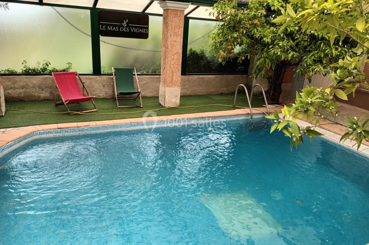 Piscine intérieure entourée de verdure avec deux chaises longues et un panneau indiquant ’Le Mas des Vignes’.