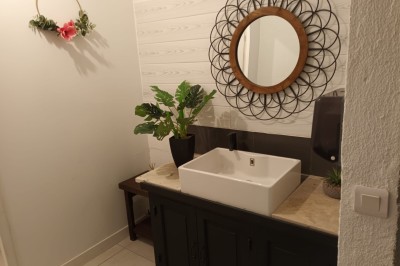 Salon lumineux avec mobilier en résine tressée, plantes vertes, tapis rond en jute et rideaux sombres.