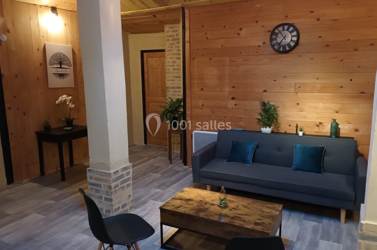 Salon chaleureux avec murs en bois, canapé gris, table basse en bois, plantes décoratives et horloge murale.