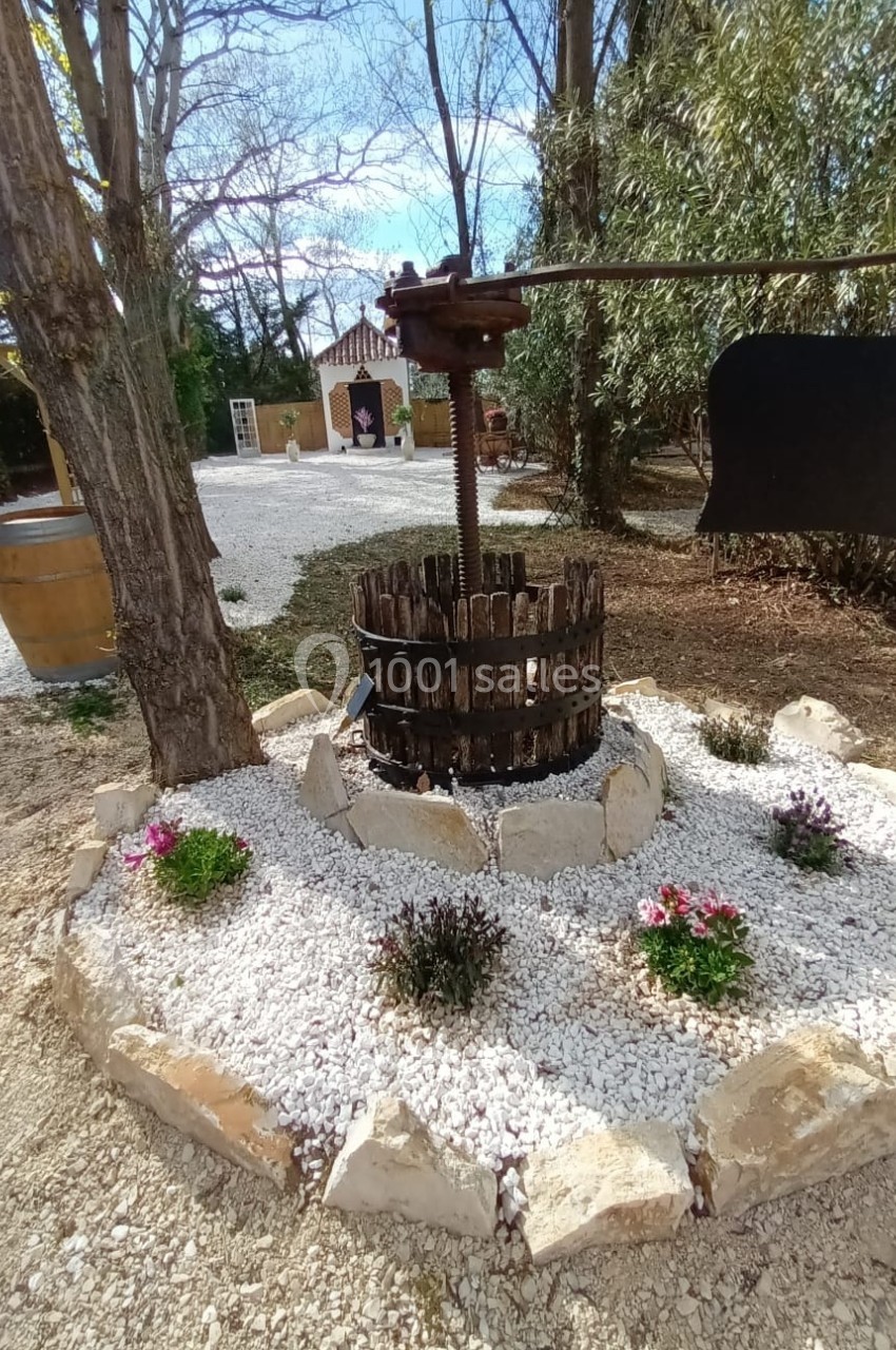 Un ancien pressoir en bois entouré de fleurs et de gravier blanc dans un jardin arboré.