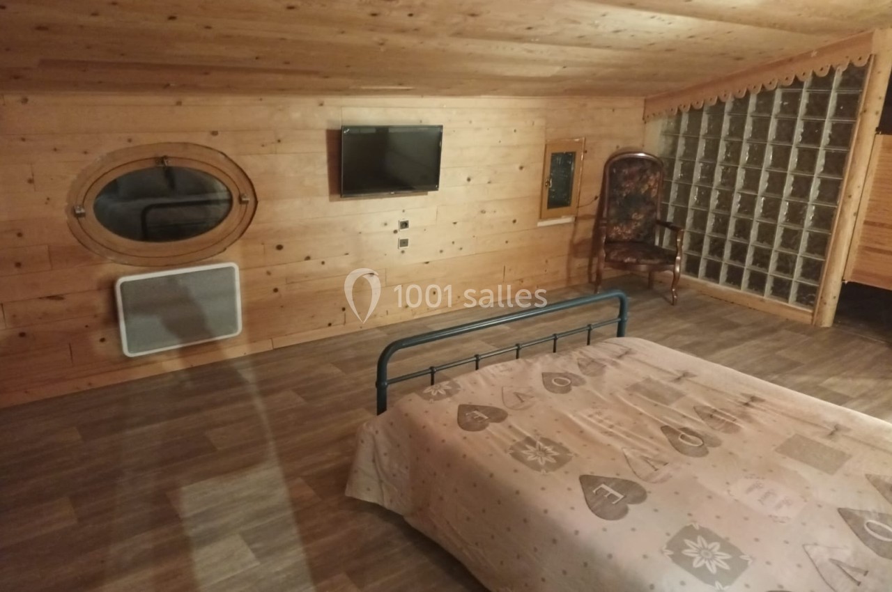 Chambre mansardée en bois avec lit double, télévision murale, fauteuil et cloison en briques de verre.