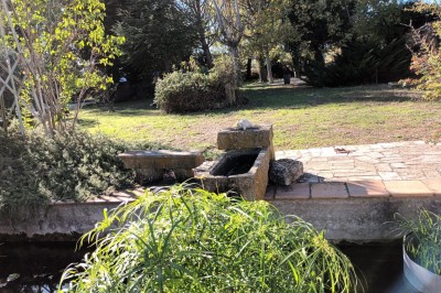 Salon lumineux avec mobilier en résine tressée, plantes vertes, tapis rond en jute et rideaux sombres.