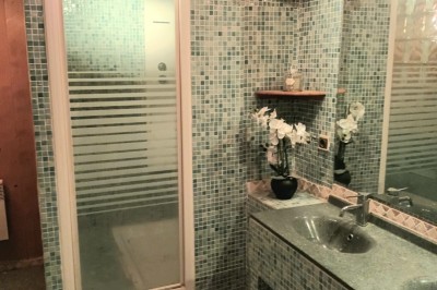 Salon lumineux avec mobilier en résine tressée, plantes vertes, tapis rond en jute et rideaux sombres.