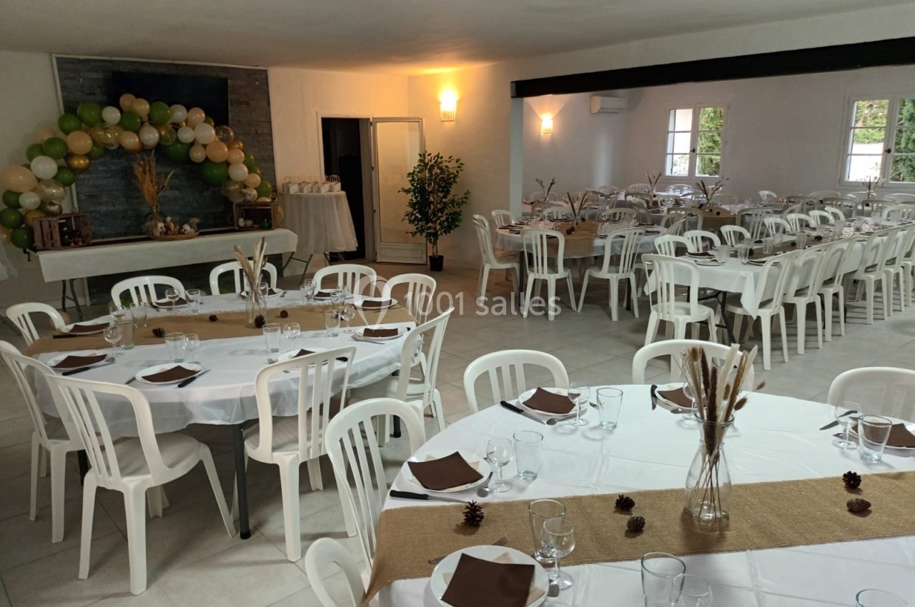 Salle décorée pour un événement avec tables rondes dressées, chaises blanches et guirlande de ballons près d'une cheminée.