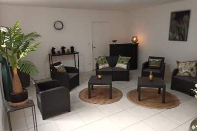 Salon lumineux avec mobilier en résine tressée, plantes vertes, tapis rond en jute et rideaux sombres.