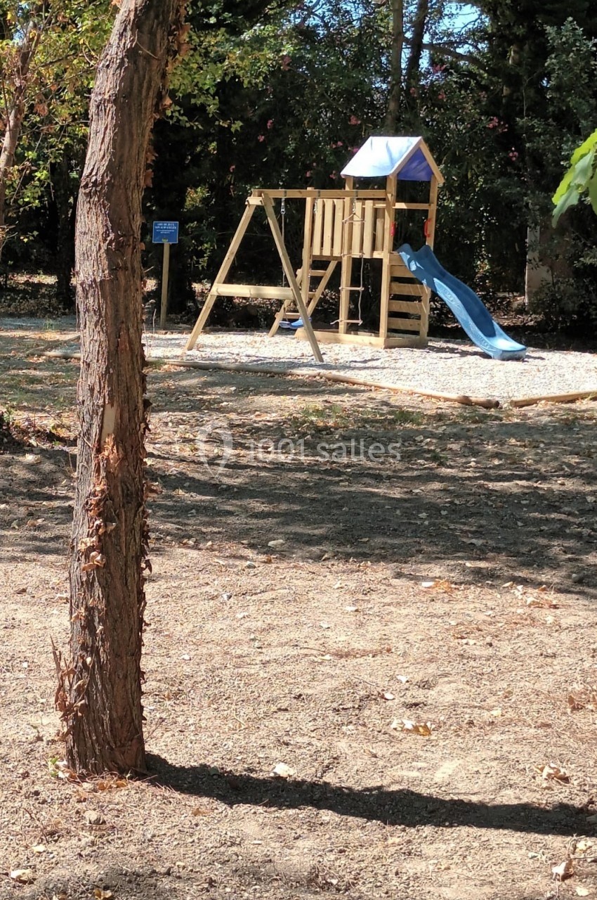 Structure de jeu en bois avec toboggan bleu et balançoires, située sur un sol gravillonné dans un espace extérieur arboré.