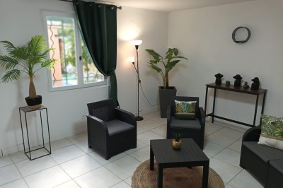 Salon lumineux avec mobilier en résine tressée, plantes vertes, tapis rond en jute et rideaux sombres.